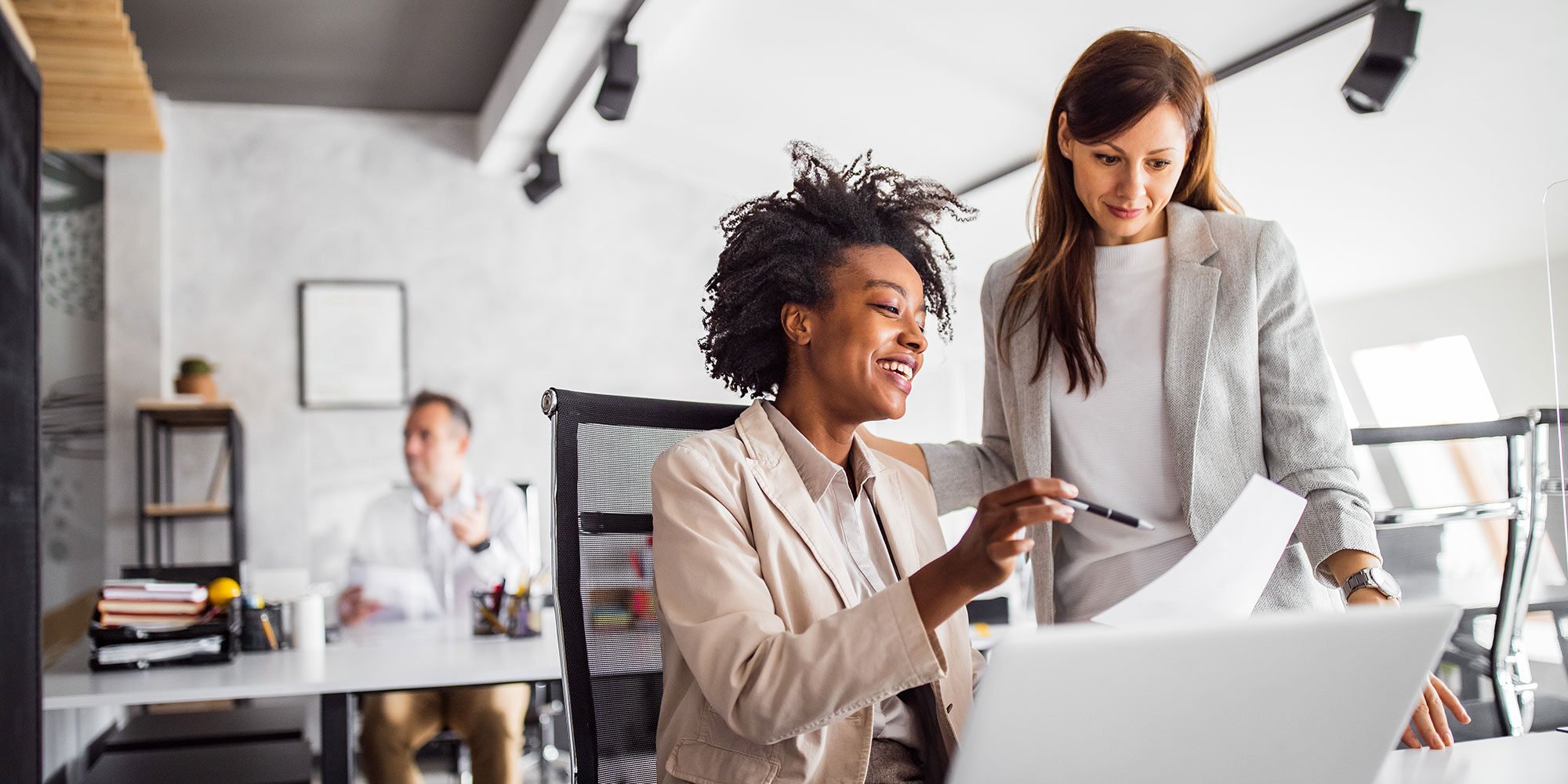 Two working women talking. One is pointing at a paper and the other is looking down at said paper. 