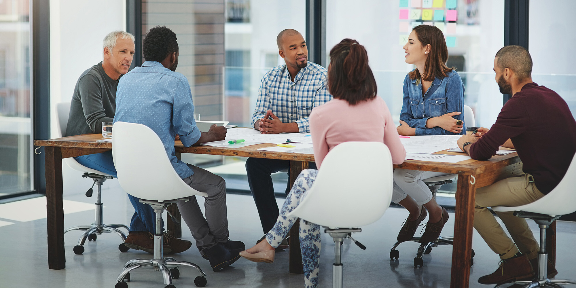 Diverse group of people engaged in a discussion around a large wooden table in a bright office space with sticky notes on the glass wall.