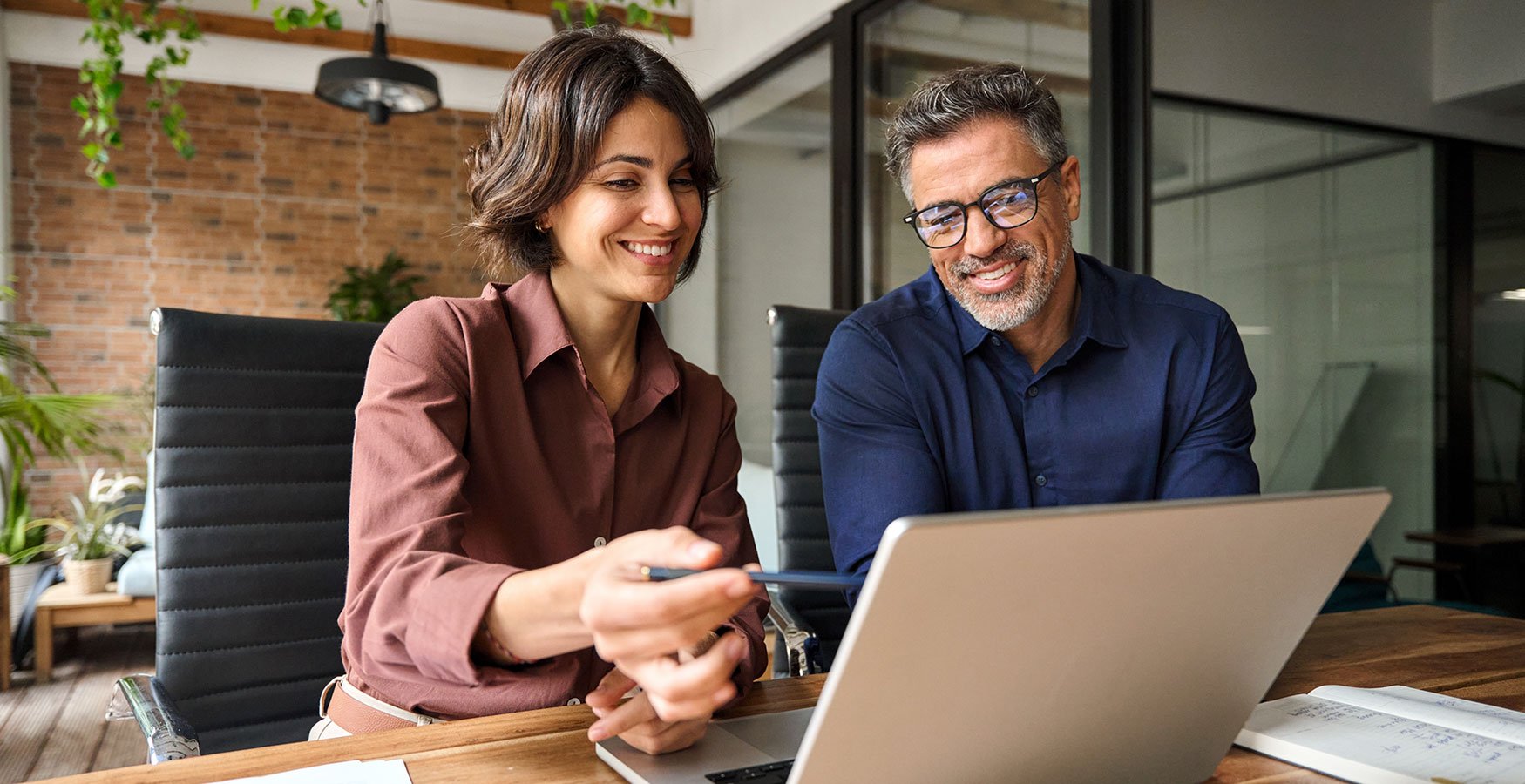 Two colleagues smiling and discussing work while looking at a laptop in a modern office.